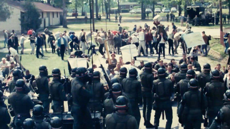 In the foreground there are police dressed in black uniforms confronting a crowd of civilians in the background on an open plain field.
