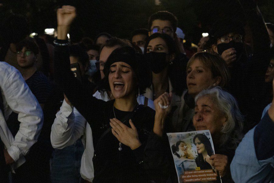 A crowd of protesters standing in Washington Square Park at night. In the center front is a woman dressed in black with her eyes closed and hand raised.