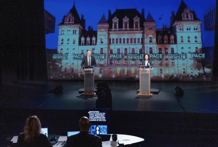 The candidates stand behind podiums on a stage. Governor Kathy Hochul is on the right talking into a microphone and U.S. representative Lee Zeldin on the left talking into a microphone.