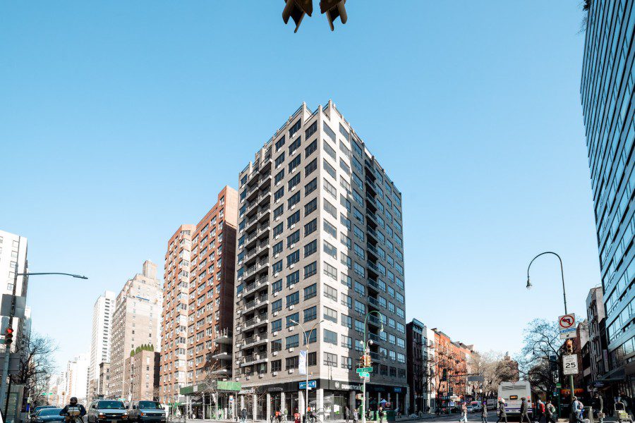 Anexteriorviewof the Coral Tower residence hall. The building has a modern white facade. On each side of the building are rows of rectangular windows and balconies. At the bottom is a black sign for a nearby pharmacy, “Duane Reade.”