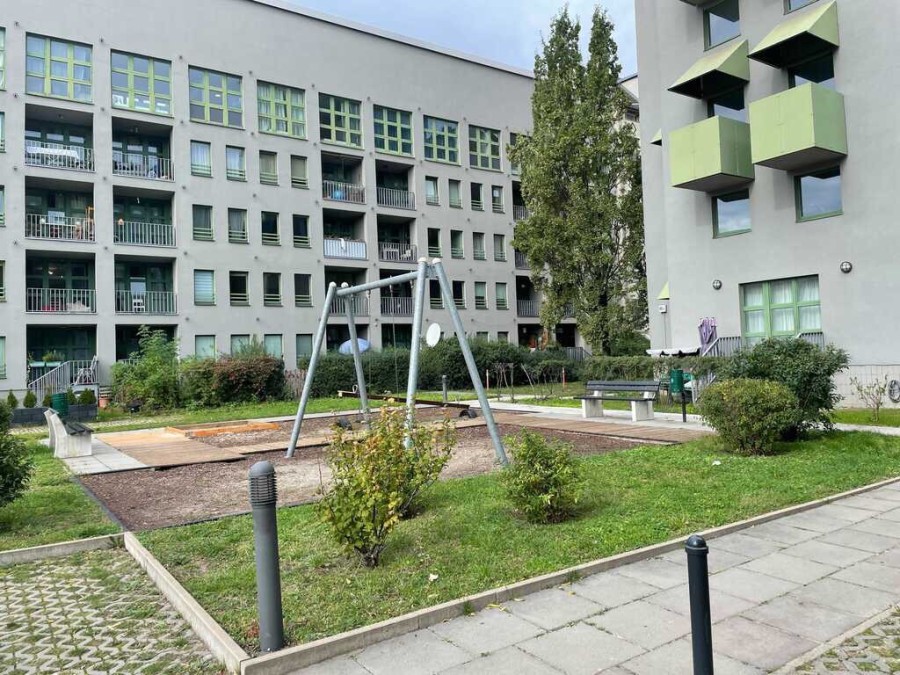 A children's playground in Berlin, with a swing set, grass and bushes. In the background, a modern building, approximately five stories tall, with rows of windows and balconies.