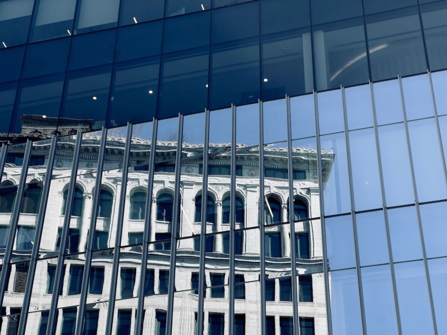 The marble facade of a pre-war building reflected in the floor-to-ceiling windows of a modern building.