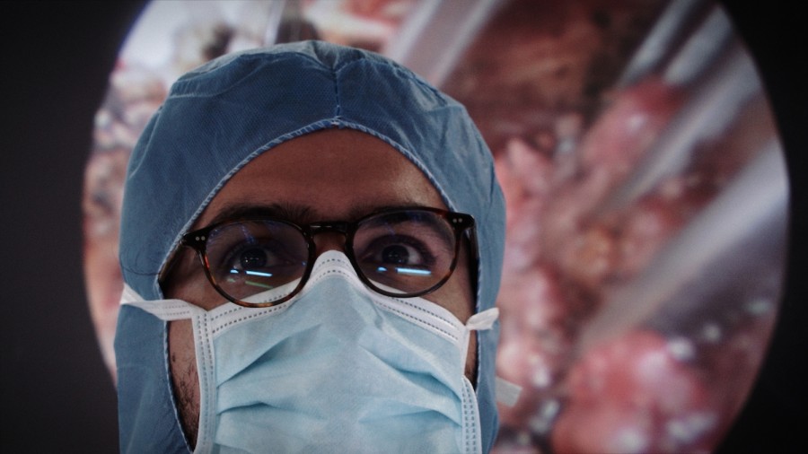 A doctor wearing a pair of black-framed glasses, a blue mask and scrubs.