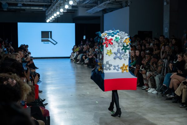 NYFWGlobalFashion-4 A model walking down a runway with an oversized blue-and-red hat with fabric flowers.