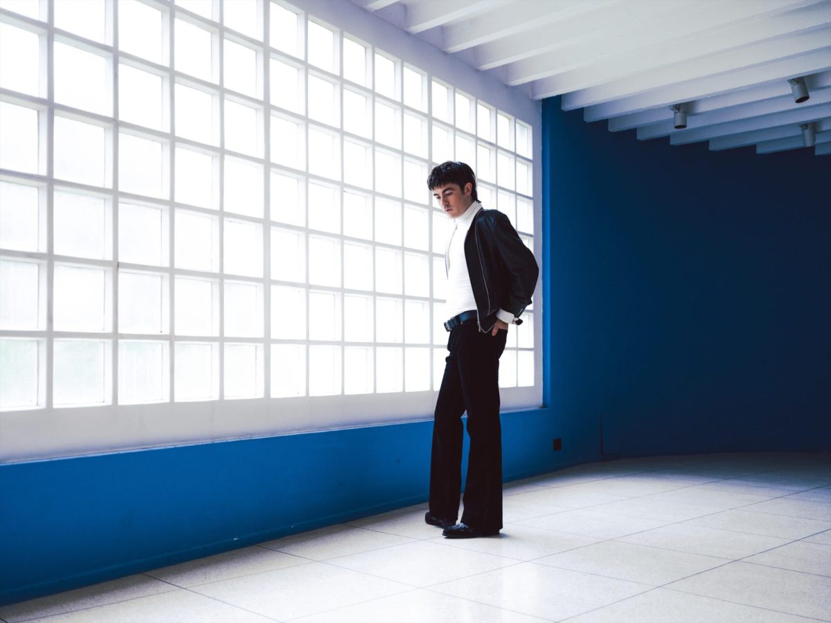 A man in a black suit standing in front of a large window with panels and a blue wall.
