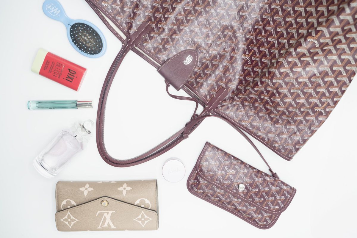 A brown bag lays flat on a white surface. Around it are a small blue hairbrush, a blush stick, a perfume rollerball, a perfume bottle, a wallet and a small jar of lip mask.