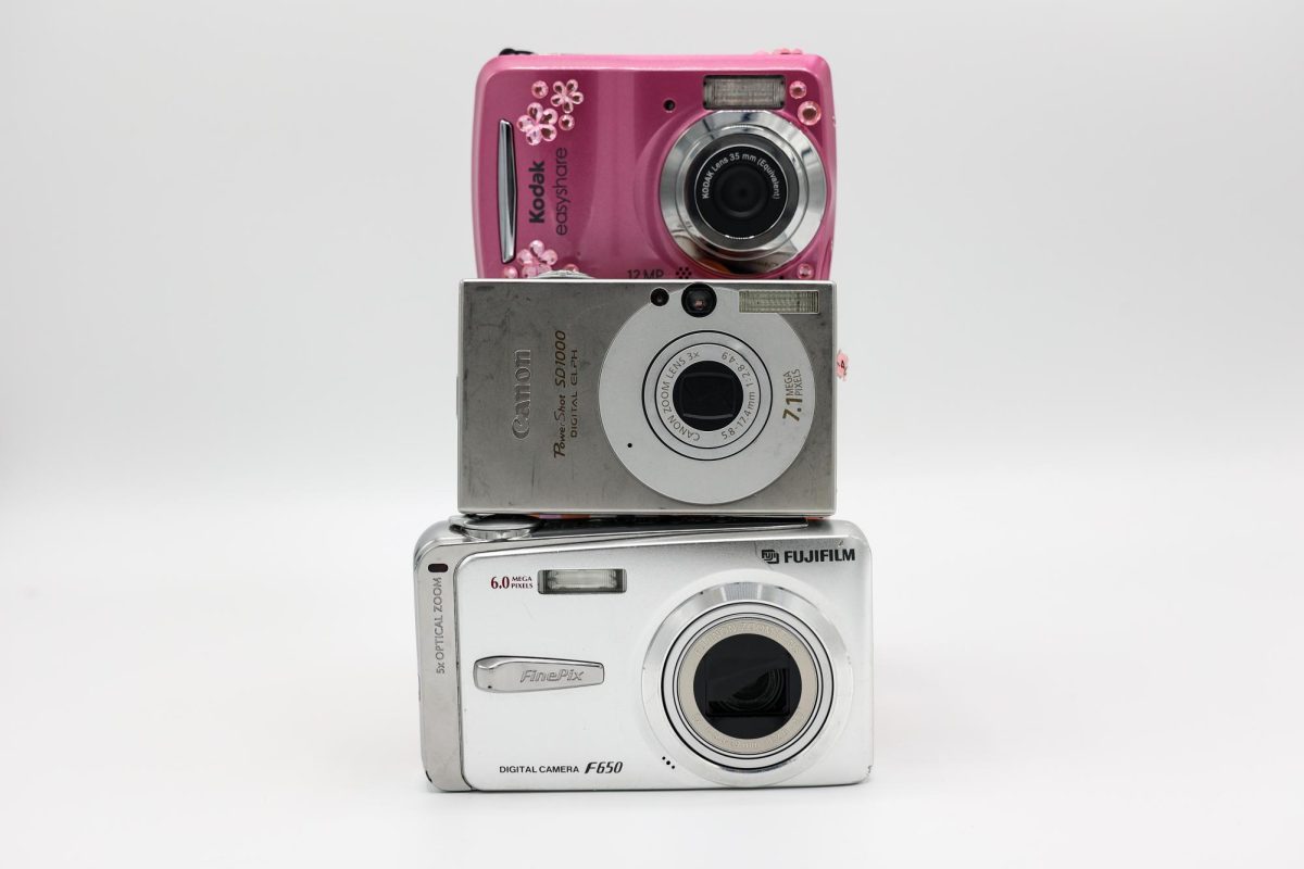Three digital cameras, two silver and one pink, stacked on top of one another.
