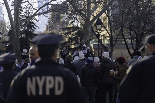 Several police officers in black caps and uniforms stand in the foreground and look on at a crowd of people in various colored outfits
